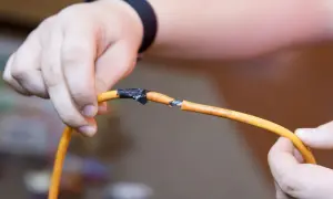 Dłonie trzymają pomarańczowy kabel z przerwaniem, pokazując, jak połączyć kable bez lutowania za pomocą taśmy izolacyjnej.