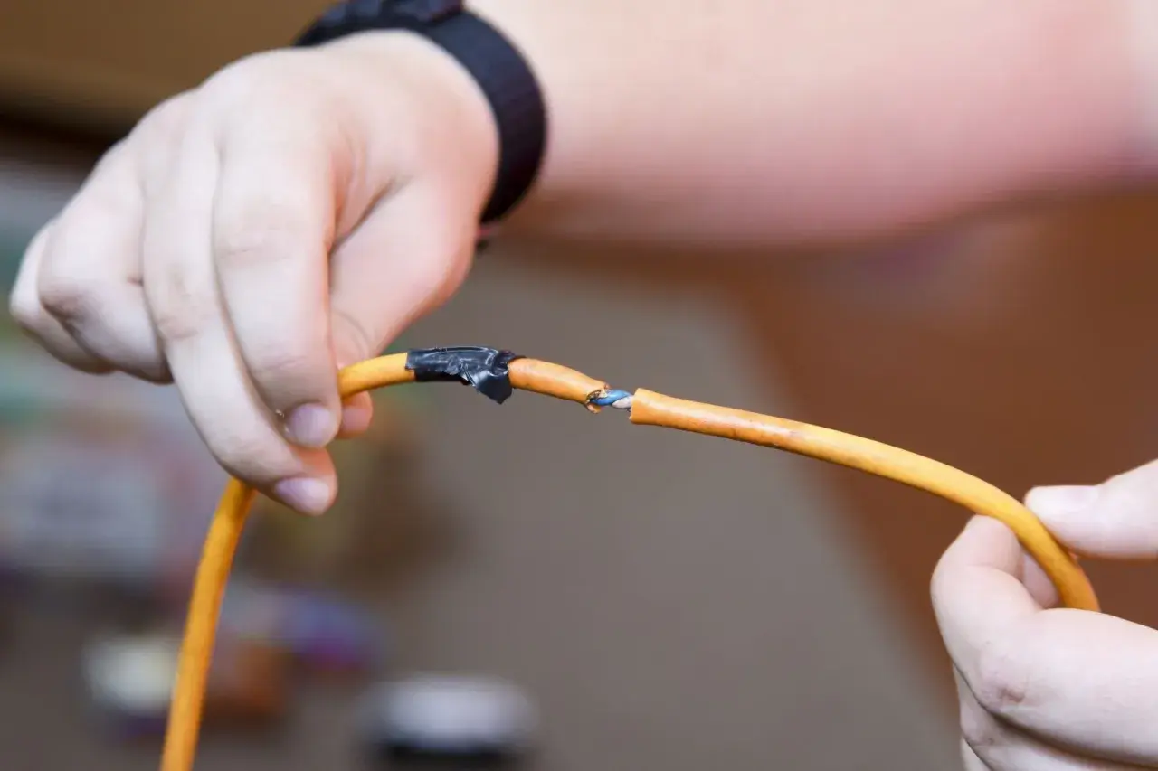 Dłonie trzymają pomarańczowy kabel z przerwaniem, pokazując, jak połączyć kable bez lutowania za pomocą taśmy izolacyjnej.